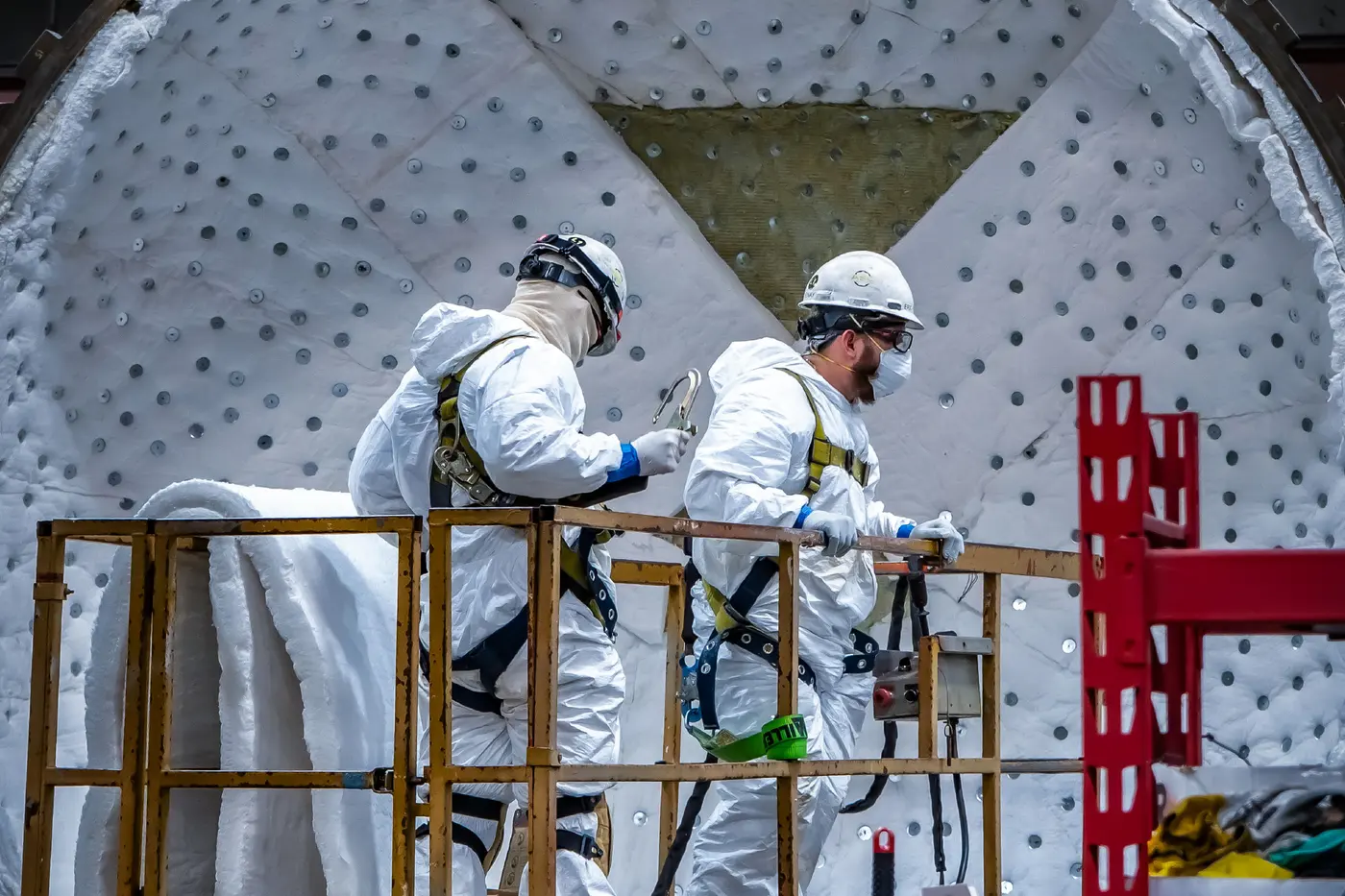 Two ASC field technicians in protective suits and harnesses installing insulation inside a large autoclave