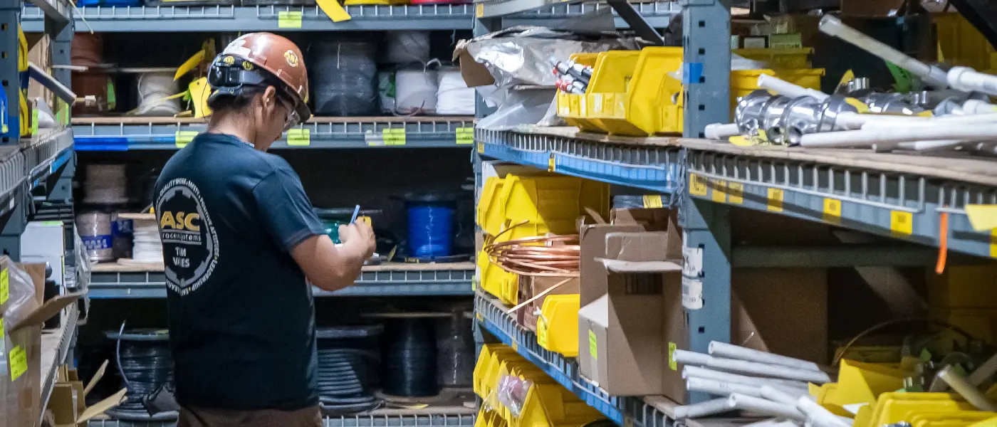 An ASC inventory clerk in a hard hat checking parts on warehouse shelves